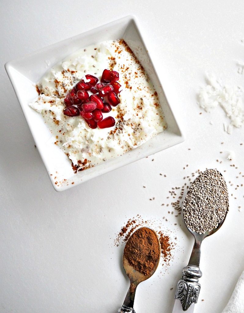 A square white bowl sits with greek yoghurt in it, topped with pomegranate seeds, chia seeds, cinnamon and grated coconut. A  spoon of chia seeds, spoon of cinnamon and some scattered coconut shavings are covering the table.