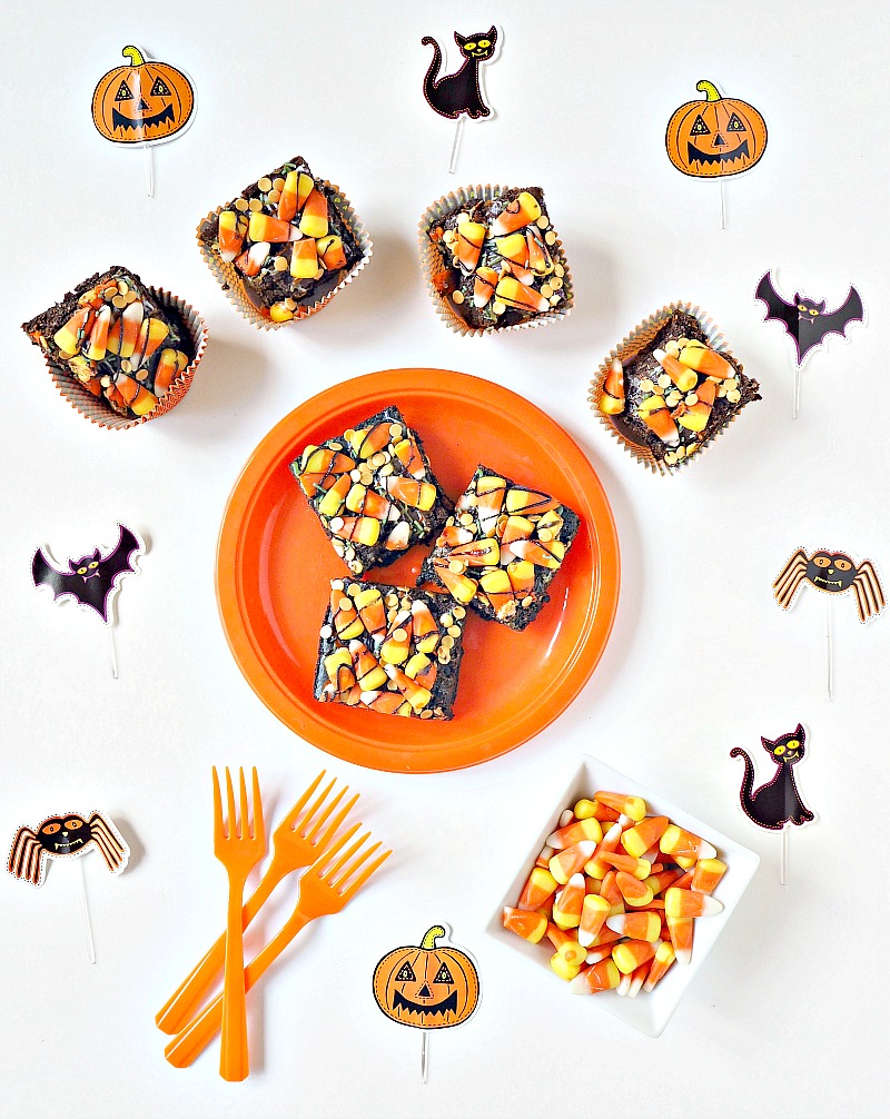 A plate of Halloween brownies surrounded by Halloween decorations and more candy corn brownies