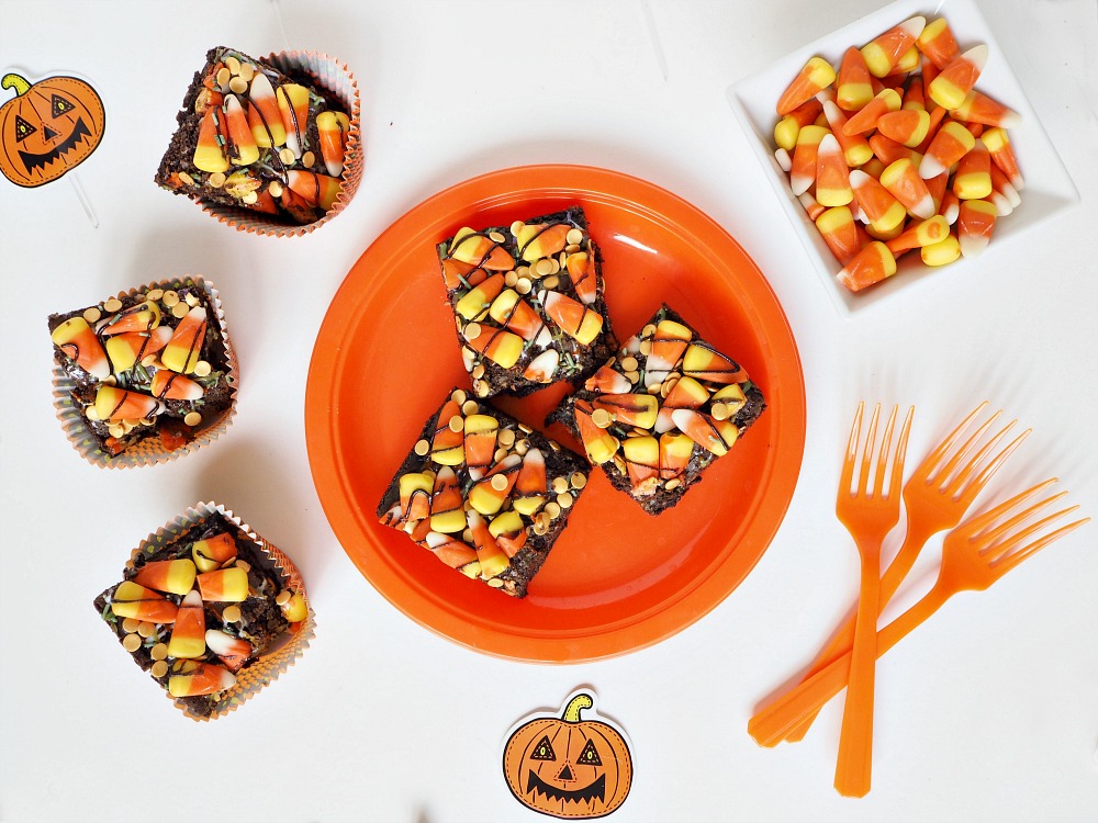 3 Halloween chocolate brownies on an orange plate, with 3 more brownies beside it, a bowl of candy corn and 3 orange forks. 