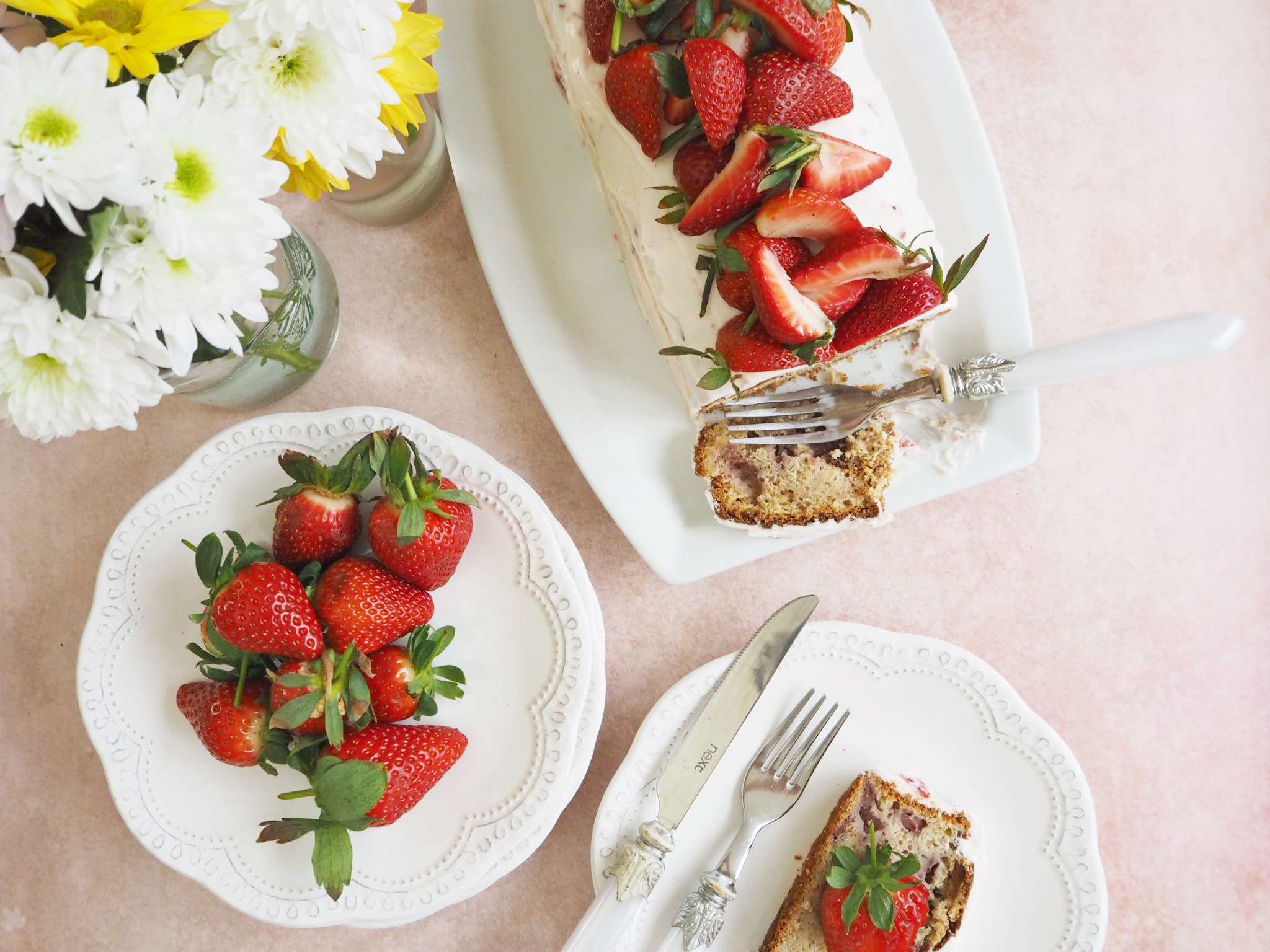 Strawberry bread with cream cheese frosting and fresh strawberries on top, sliced to show the inside of the loaf. A slice is on a plate to the right of the bread, and another plate has additional strawberries