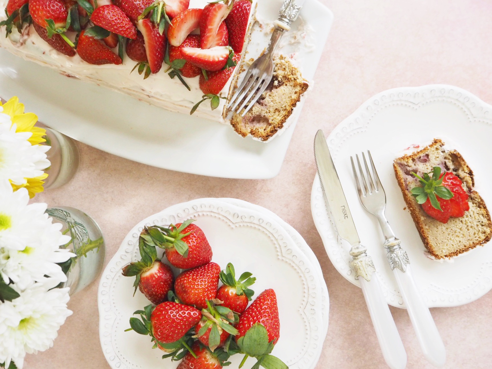 Strawberry bread with cream cheese frosting and fresh strawberries on top, sliced to show the inside of the loaf. A slice is on a plate to the right of the bread, and another plate has additional strawberries