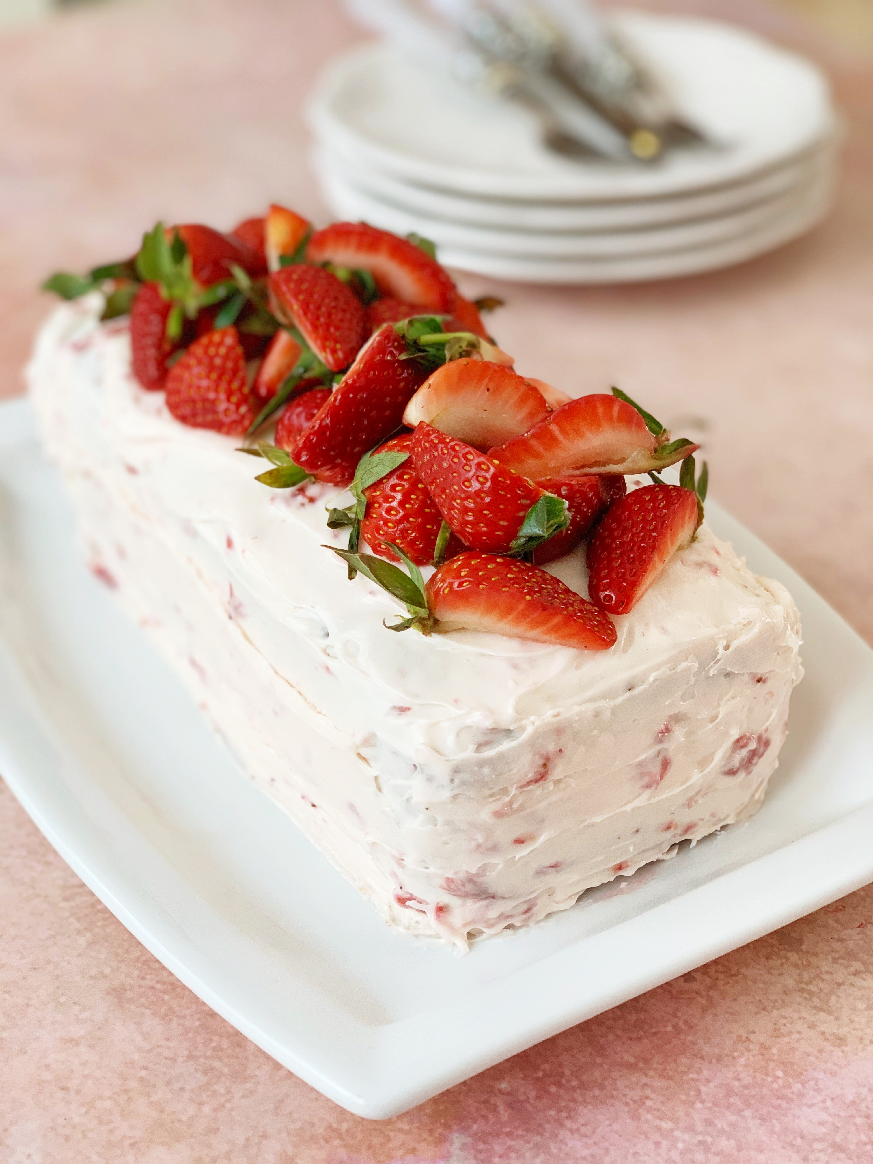 Strawberry bread with cream cheese frosting and fresh strawberries, with a stack of plates out of focus in the background