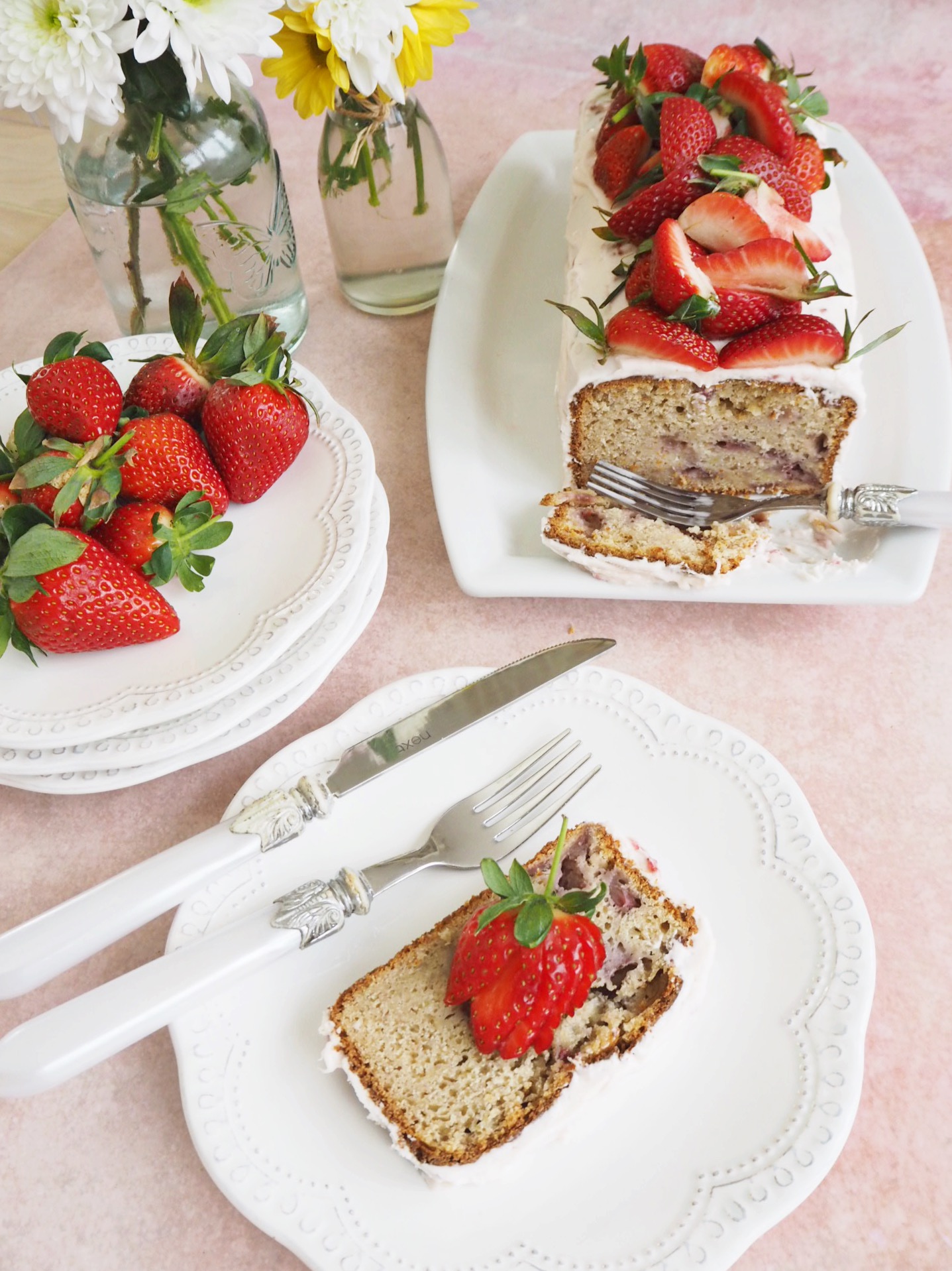 Strawberry bread with cream cheese frosting and fresh strawberries on top, sliced to show the inside of the loaf. A slice is on a plate to the right of the bread, and another plate has additional strawberries