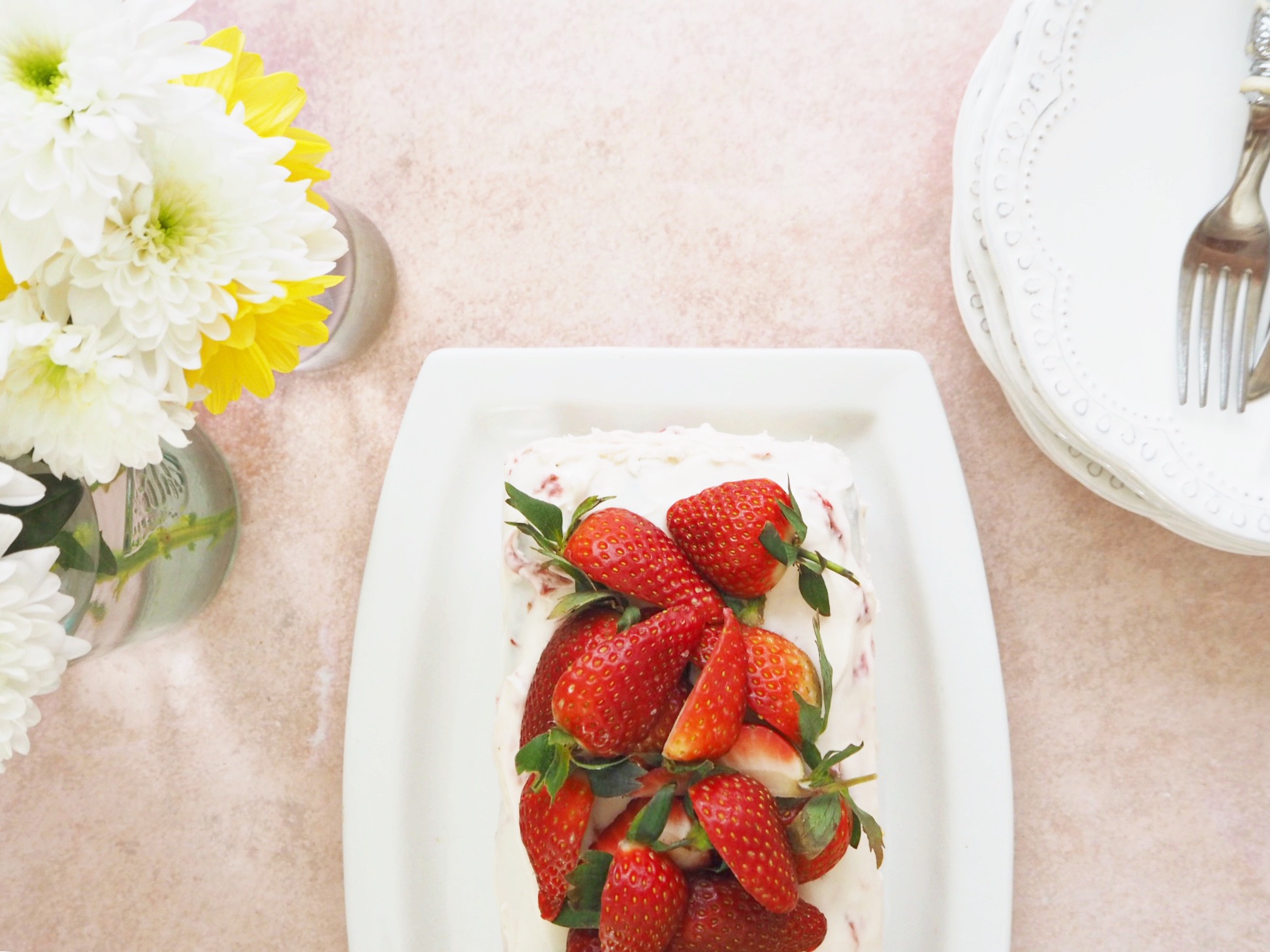 Strawberry bread with cream cheese frosting and fresh strawberries on top. To the left of it is a bunch of flowers in spring colours