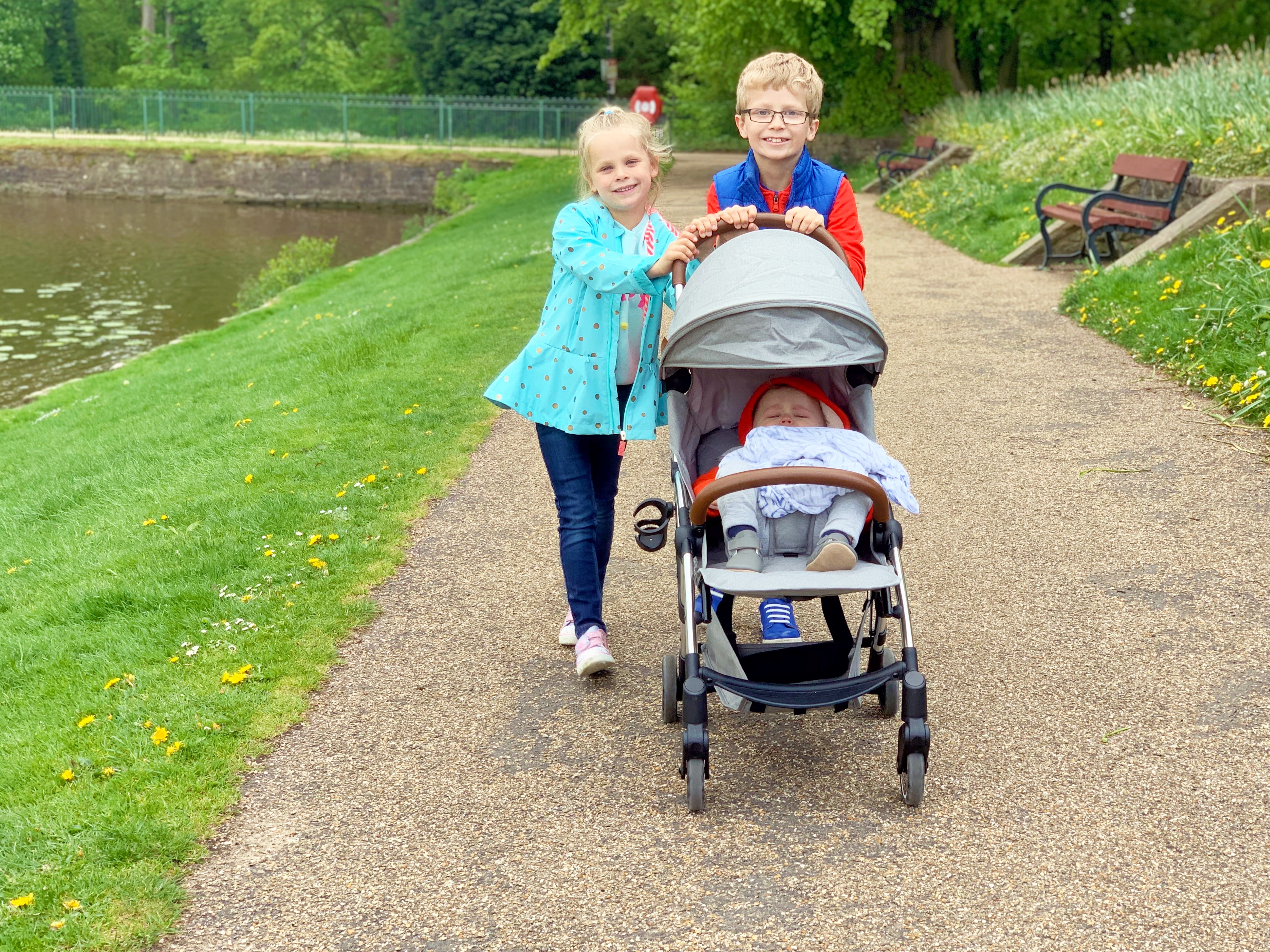 A brother and sister push their baby brother in a grey stroller while he sleeps in the park