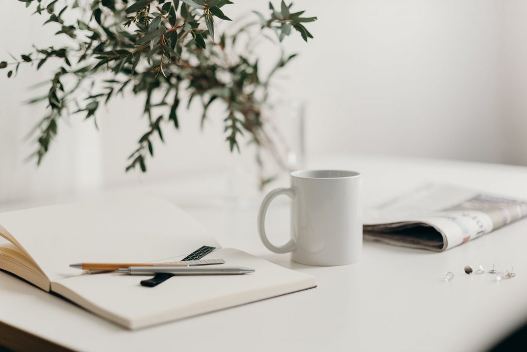 A white desk with an open notebook, pens, a white mug and a leafy plant on it