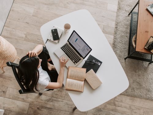 A bird's eye view of a woman sitting at a white desk. She is working on a laptop with notebooks around the desk