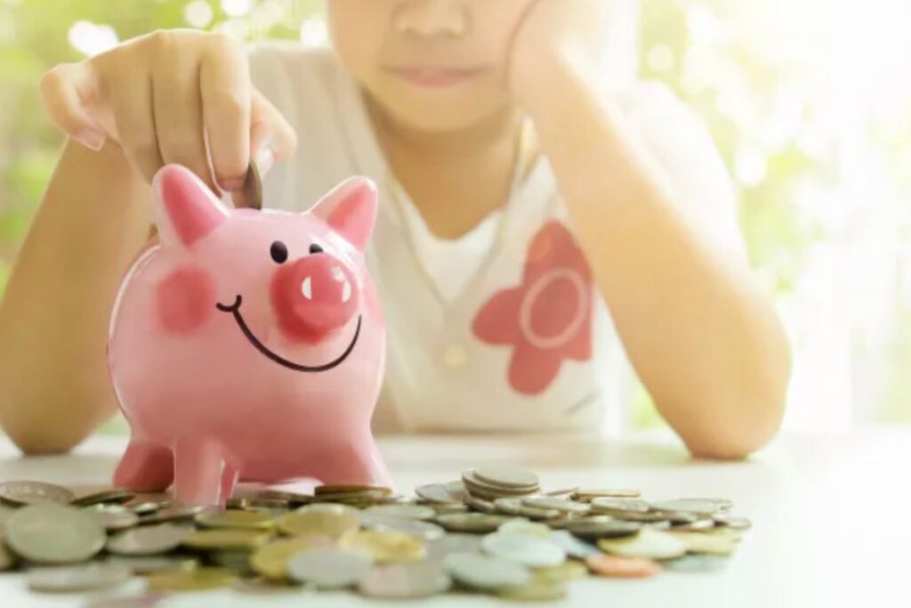 A Piggy bank shaped like a pink sits on a table, surrounded by coins. A child is placing a coin into the bakc of the piggy bank