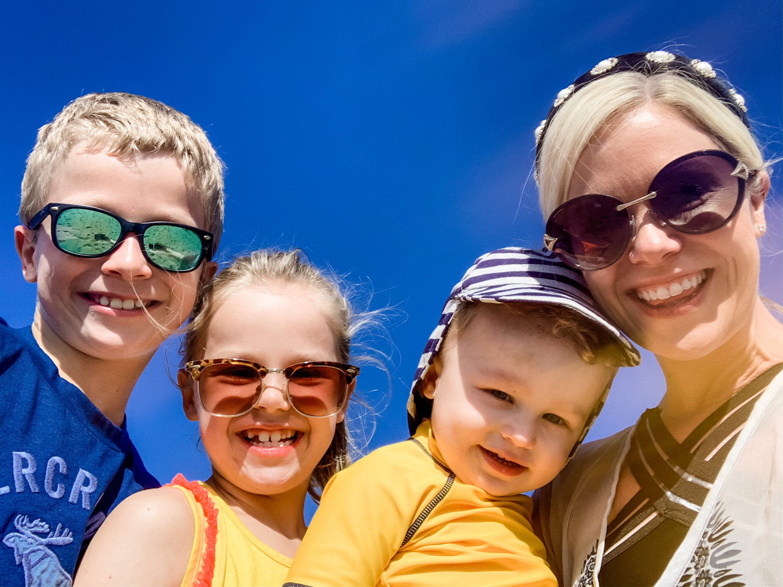 A mum and 3 children smiling for the camera with a bright blue sky behind them. 3 of them are wearing sunglasses and the baby is wearing a hat.