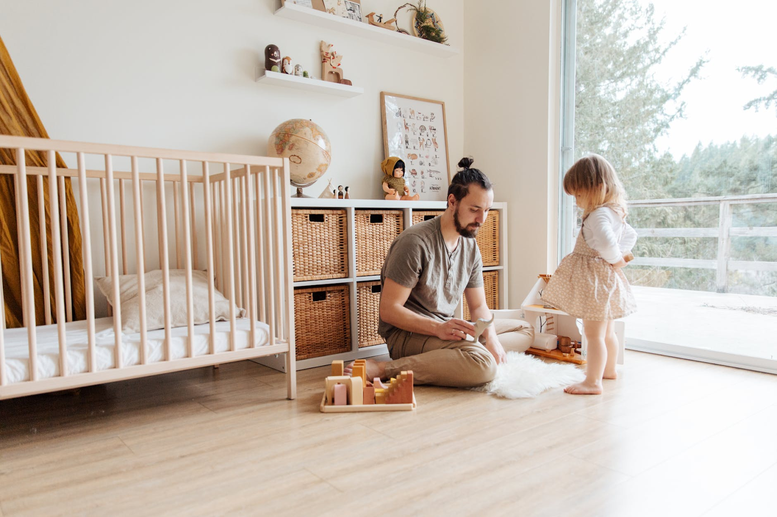 A man sits on the floor of a neutral coloured nursery and plays with a young girl standing in front of him