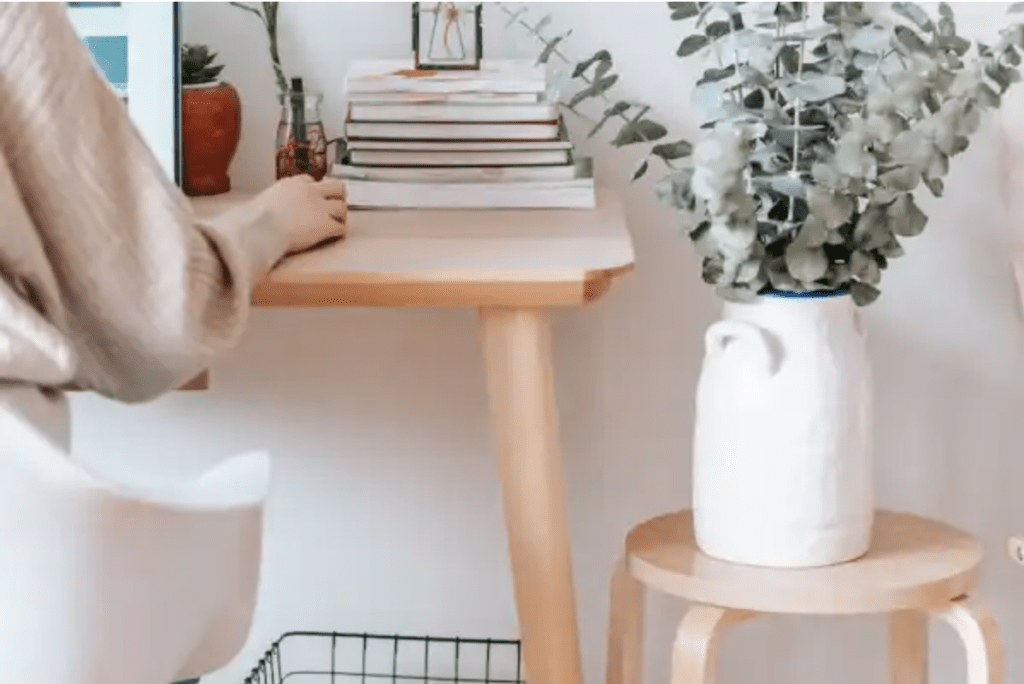 A desk with a person sitting at it. A stool next to the desk has a vase and a plant on it