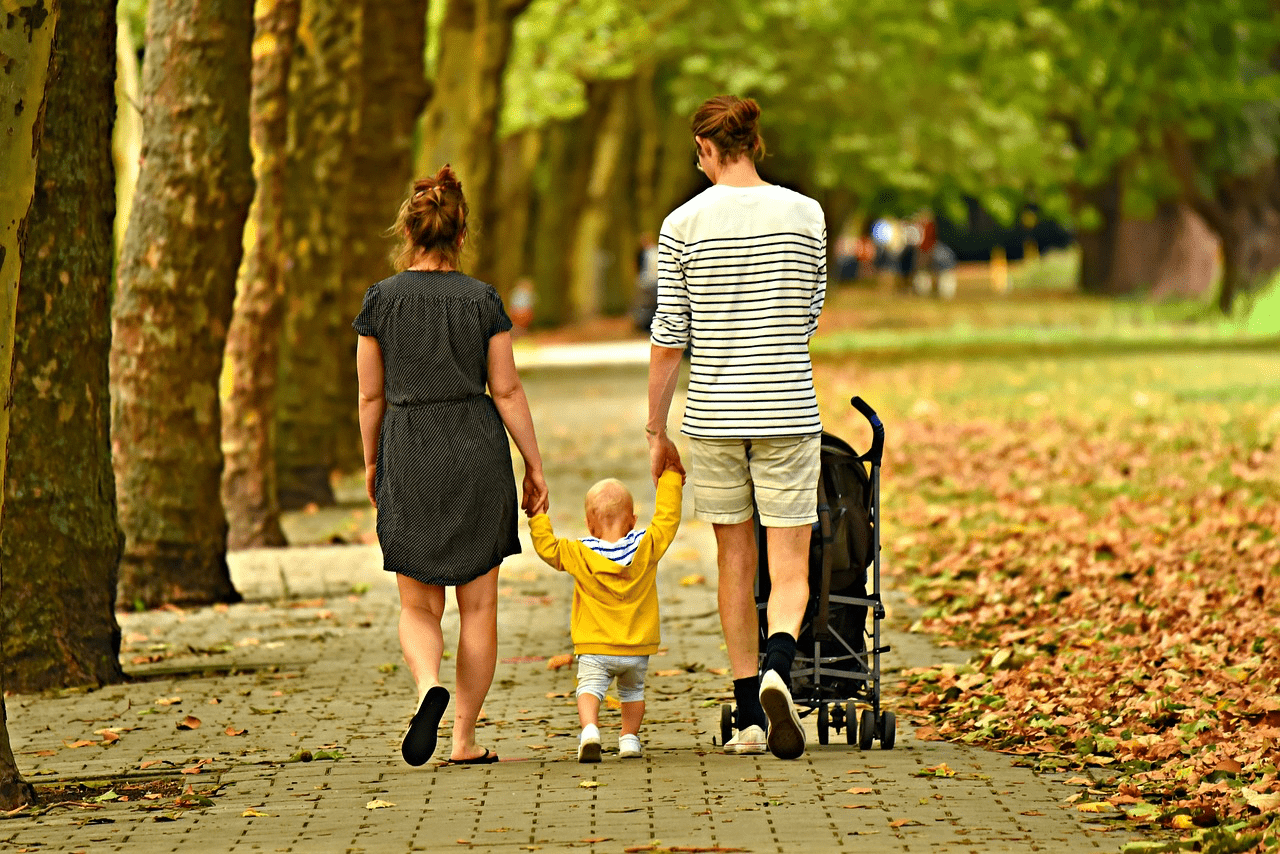 A man and woman walking down a path, holding the hands of a toddler between them. There are trees lining one side of the path and grass and fallen leaves on the other