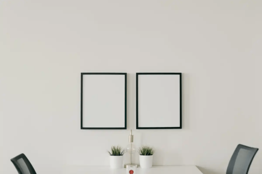 A white wall with two black frames on it. At the bottom of the picture you can see a white tables with 2 chairs and two small plants