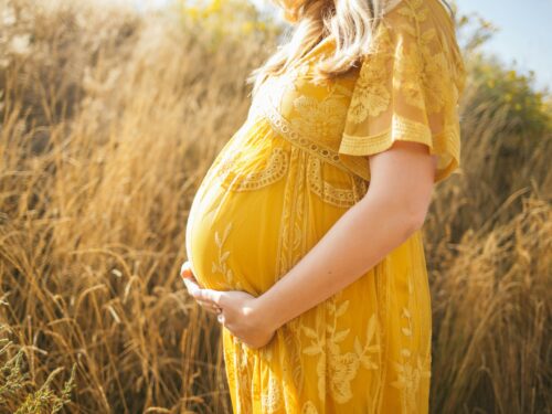 A pregnant woman in a yellow dress gracefully balancing motherhood while standing in a field, prioritizing her prenatal health.