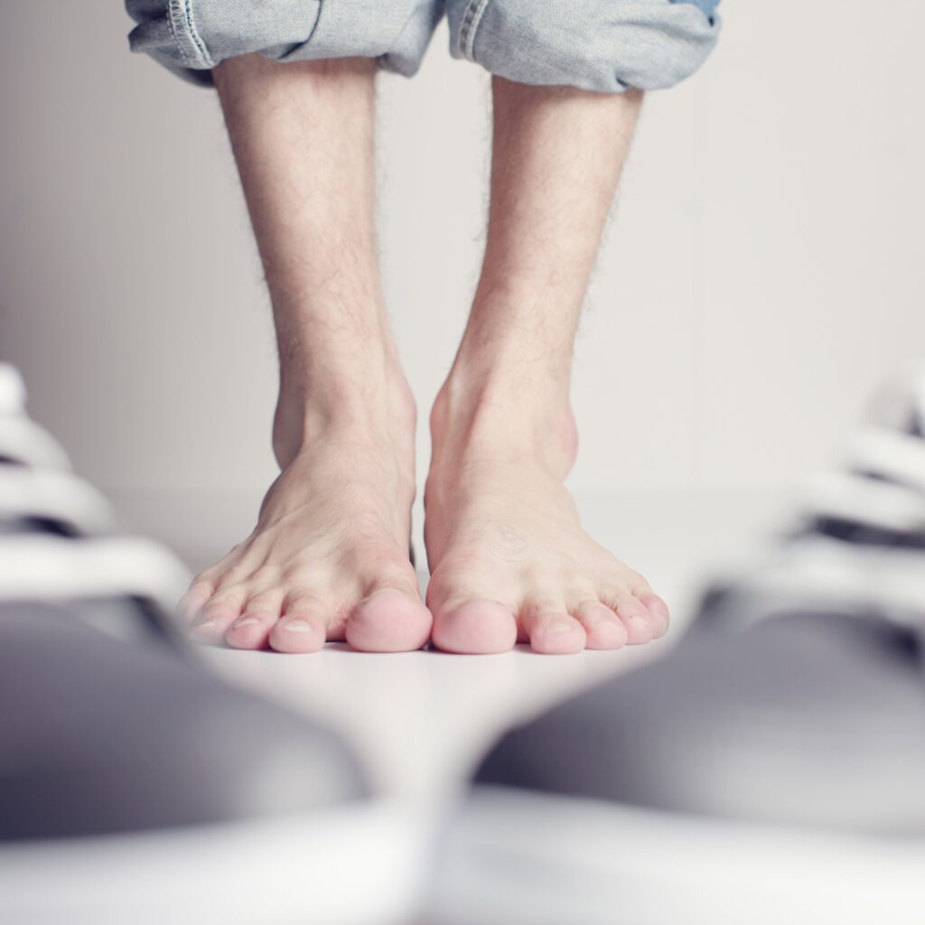 A man's bare feet standing next to a pair of sneakers, showing signs of the need for arch support.