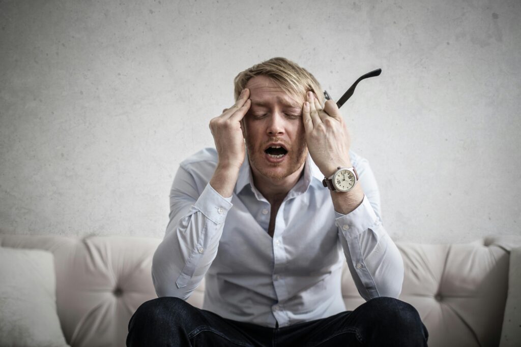 A man sitting on a couch with his hands on his head, expressing chronic pain.