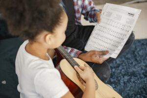 A young girl is learning music at an early age as she plays an acoustic guitar with her father, displaying discipline.