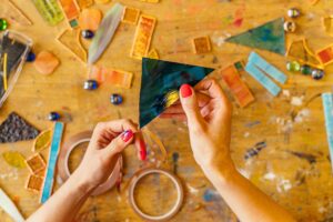 A person's hands holding a piece of colored glass above a messy craft table with various materials for DIY Art Projects.