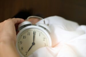 Person in bed holding an alarm clock showing 7 o'clock, trying to sleep off the effects of alcohol.