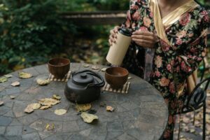 A person in a floral robe pouring a beverage from a thermal flask into a cup near a traditional teapot and cups on an outdoor table with scattered leaves, embodying Japanese aesthetics.