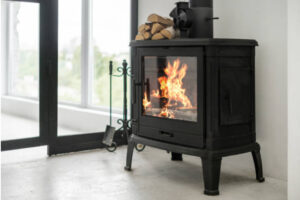 A wood-burning stove with visible fire, accompanied by a stack of logs and fireplace tools, embodies rustic radiance in a room with a white interior and a large window.