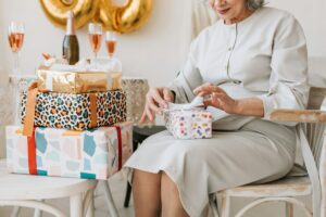 An elderly woman opening an heirloom gift at a celebration with balloons and presents in the background.